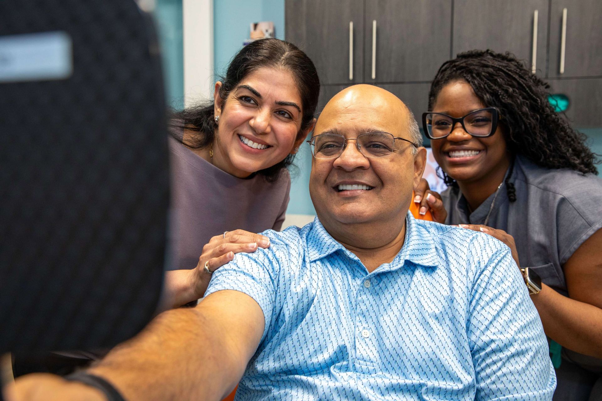 A patient smiles into a hand mirror beside Dr. Uttma Dham and a team member